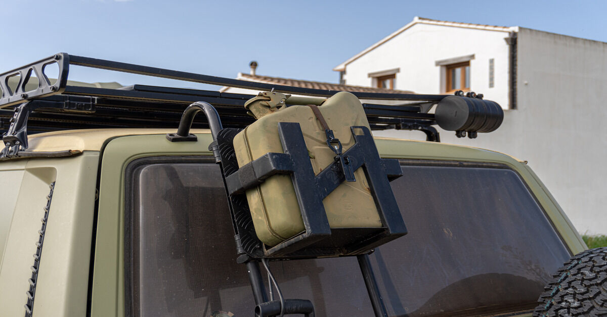 A brownish-green metal jerry can attached to the outside roof of an off-road vehicle with a jerry can mount.
